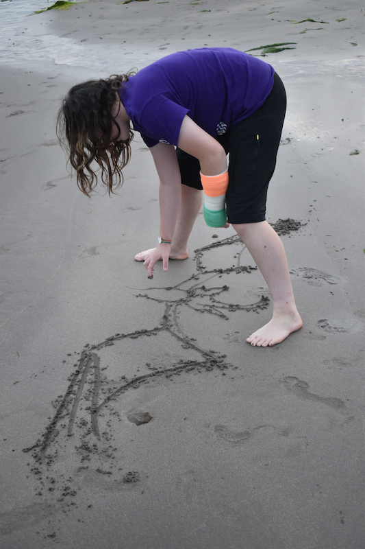 Celia drawing a dragon on a sandy beach